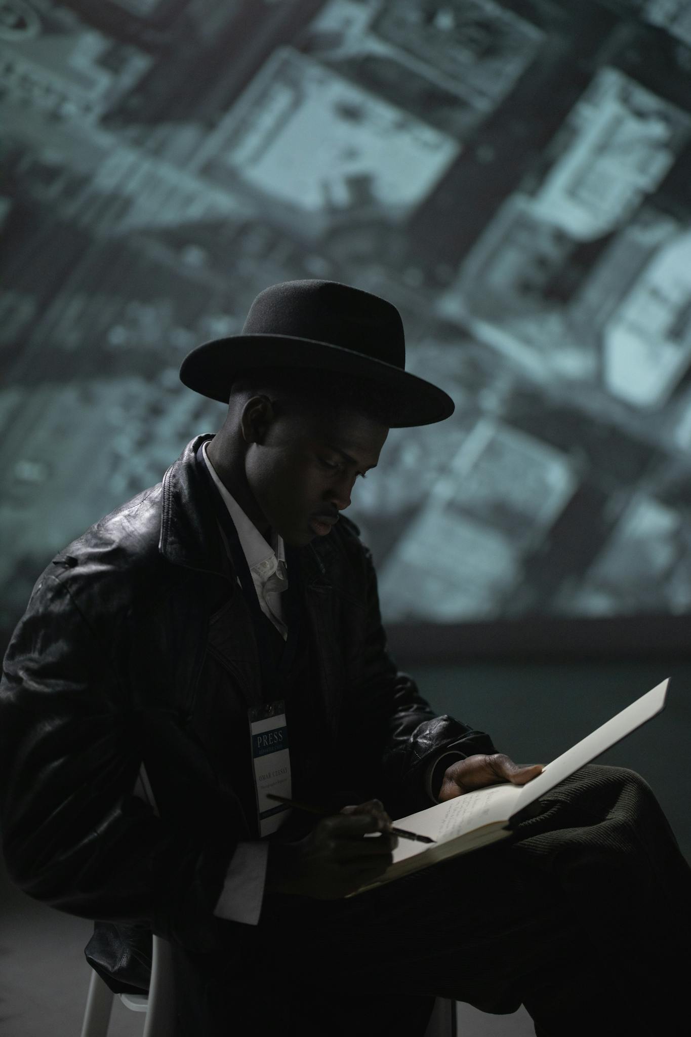 A man in a fedora and leather coat writes notes with a pen in a dimly lit studio.
