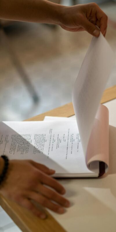 Hands flipping through a printed screenplay on a light-colored table indoors.
