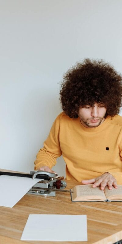 Young adult reading a book beside a typewriter, focused at a wooden table indoors.